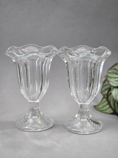 Zwei transparente Eisbecher aus Glas stehen nebeneinander auf grauem Untergrund, beide haben einen gewellten Rand, einen hohen Kelch und einen runden Fuß