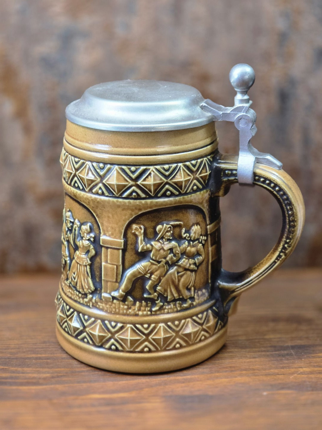 Vintage Gerzit Bierkrug aus braun glasiertem Steinzeug mit Zinndeckel, Henkel und umlaufenden Reliefs tanzender Paare in Seitenansicht.