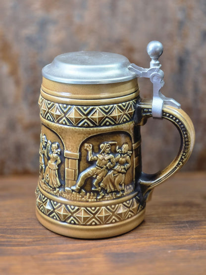 Vintage Gerzit Bierkrug aus braun glasiertem Steinzeug mit Zinndeckel, Henkel und umlaufenden Reliefs tanzender Paare in Seitenansicht.