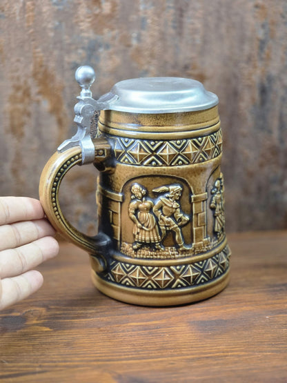 Vintage Gerzit Bierkrug mit Zinndeckel, brauner Glasur und plastischem Figurenrelief, daneben eine Hand zur Darstellung der Größe.