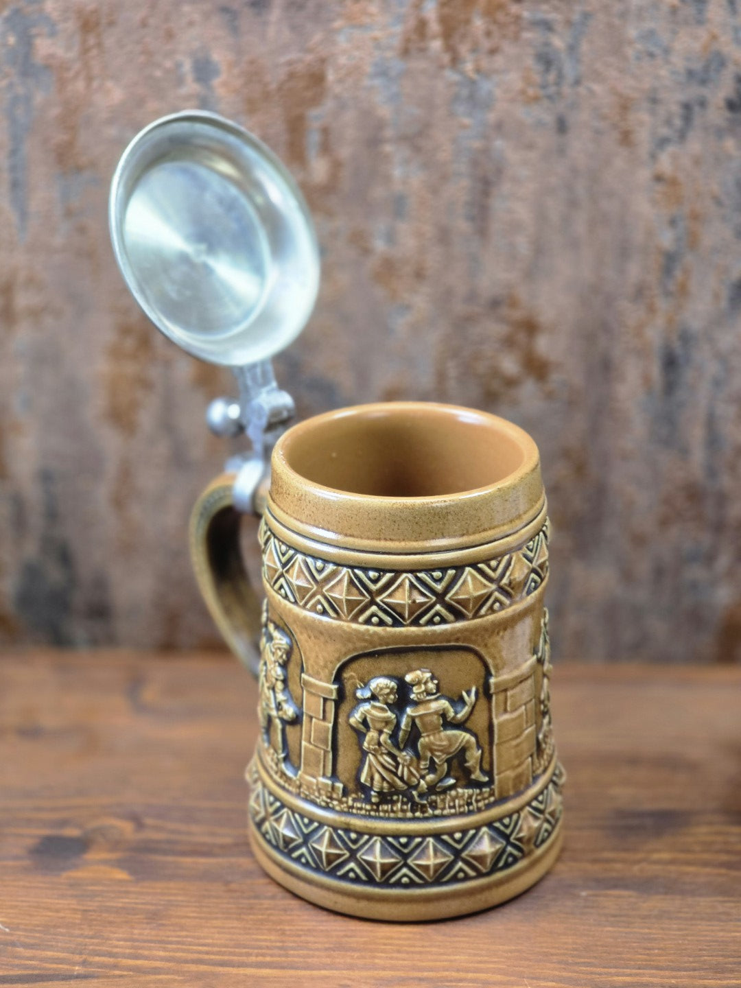 Vintage Gerzit Bierkrug mit geöffnetem Zinndeckel, braun glasierter Innenfläche und umlaufenden Figurenreliefs auf Holztisch.