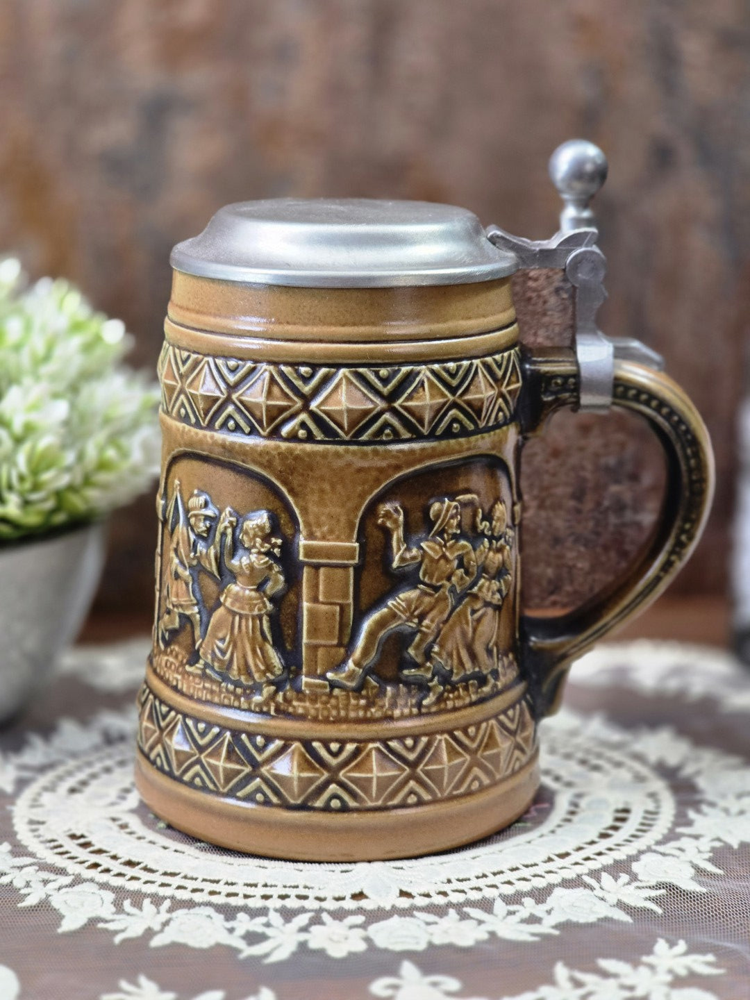 Vintage Gerzit Bierkrug mit Zinndeckel, Henkel und umlaufenden Reliefs tanzender Paare auf dekorativer Spitzendecke.