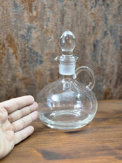 Vintage Glasdekanter mit rundem Bauch, Glasstöpsel, Henkel und Hand zum Größenvergleich auf Holztisch