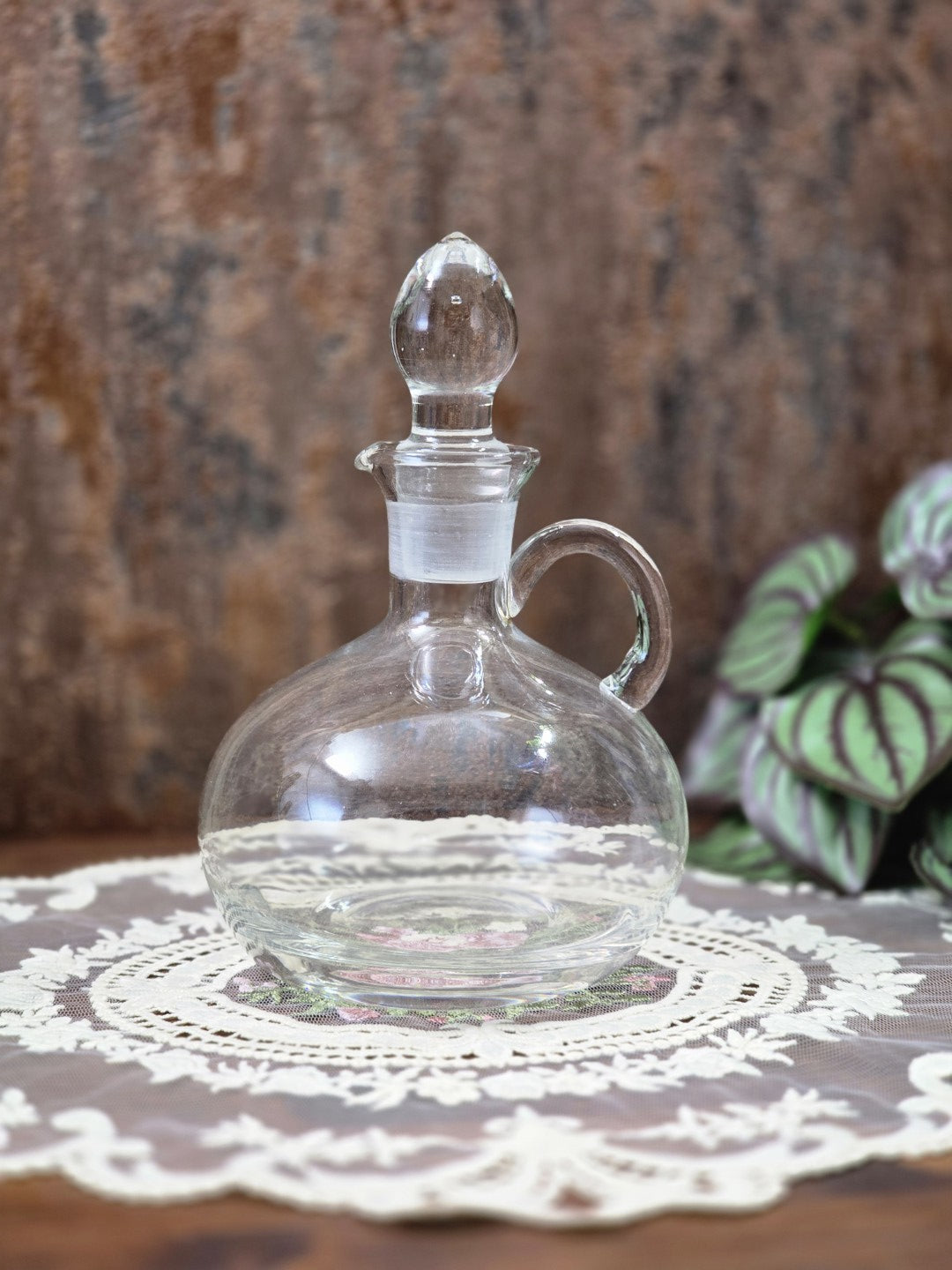 Vintage Glasdekanter mit eingesetztem Stöpsel, rundem Korpus und Henkel auf weißer Spitzendecke
