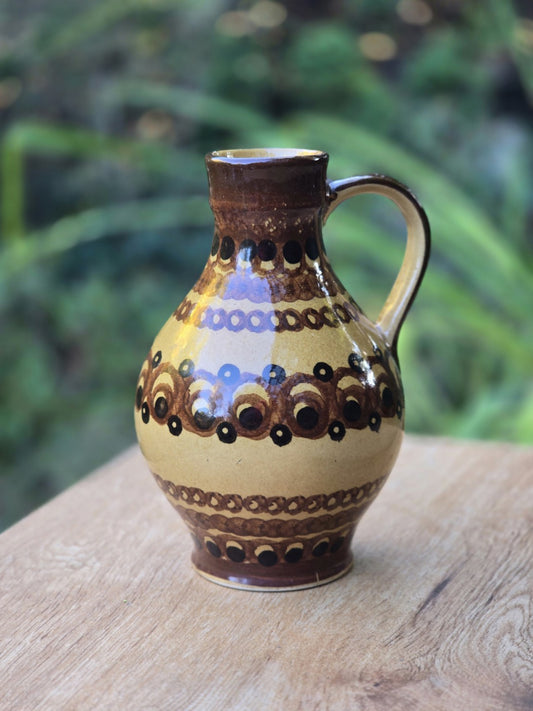 Vintage Keramik-Henkelkrug mit bauchiger Form und geometrischem Dekor in Ockergelb, Braun und Schwarz auf hellem Holztisch.