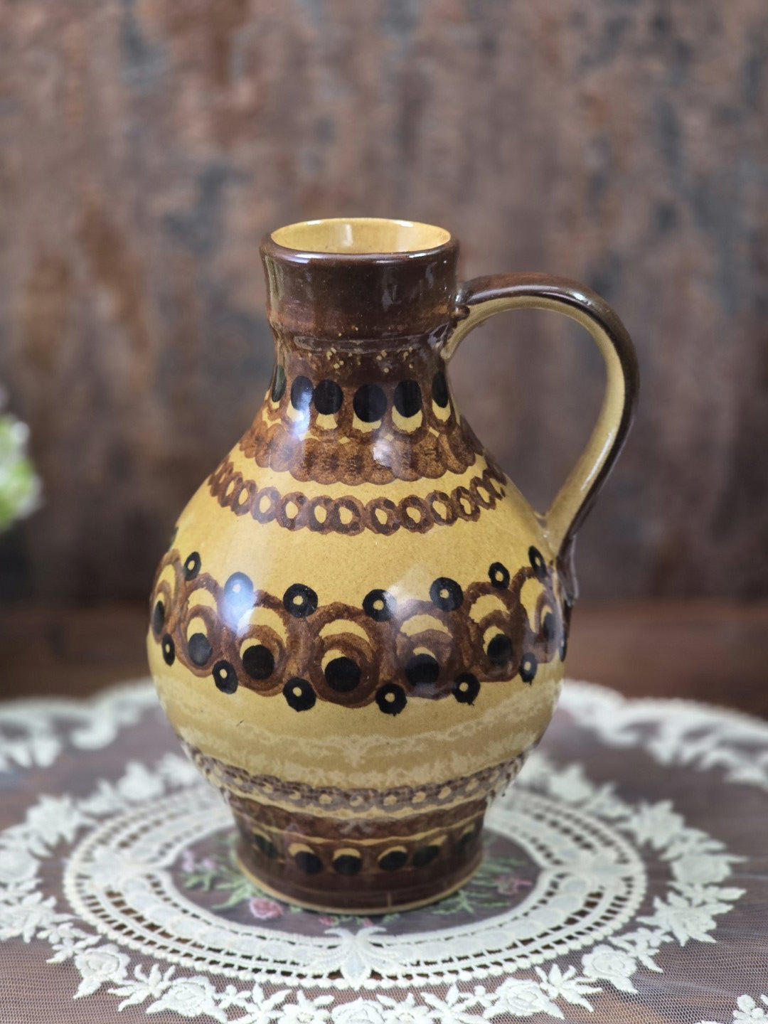 Vintage Keramik-Henkelkrug mit bauchigem Korpus und ornamentalen Bändern in Ockergelb, Braun und Schwarz auf weißer Spitzendecke.