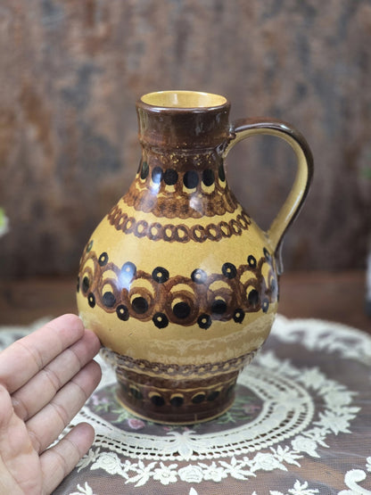 Vintage Keramikkrug mit braunem Henkel und geometrischem Dekor in Ocker und Schwarz neben einer Hand als Größenvergleich.