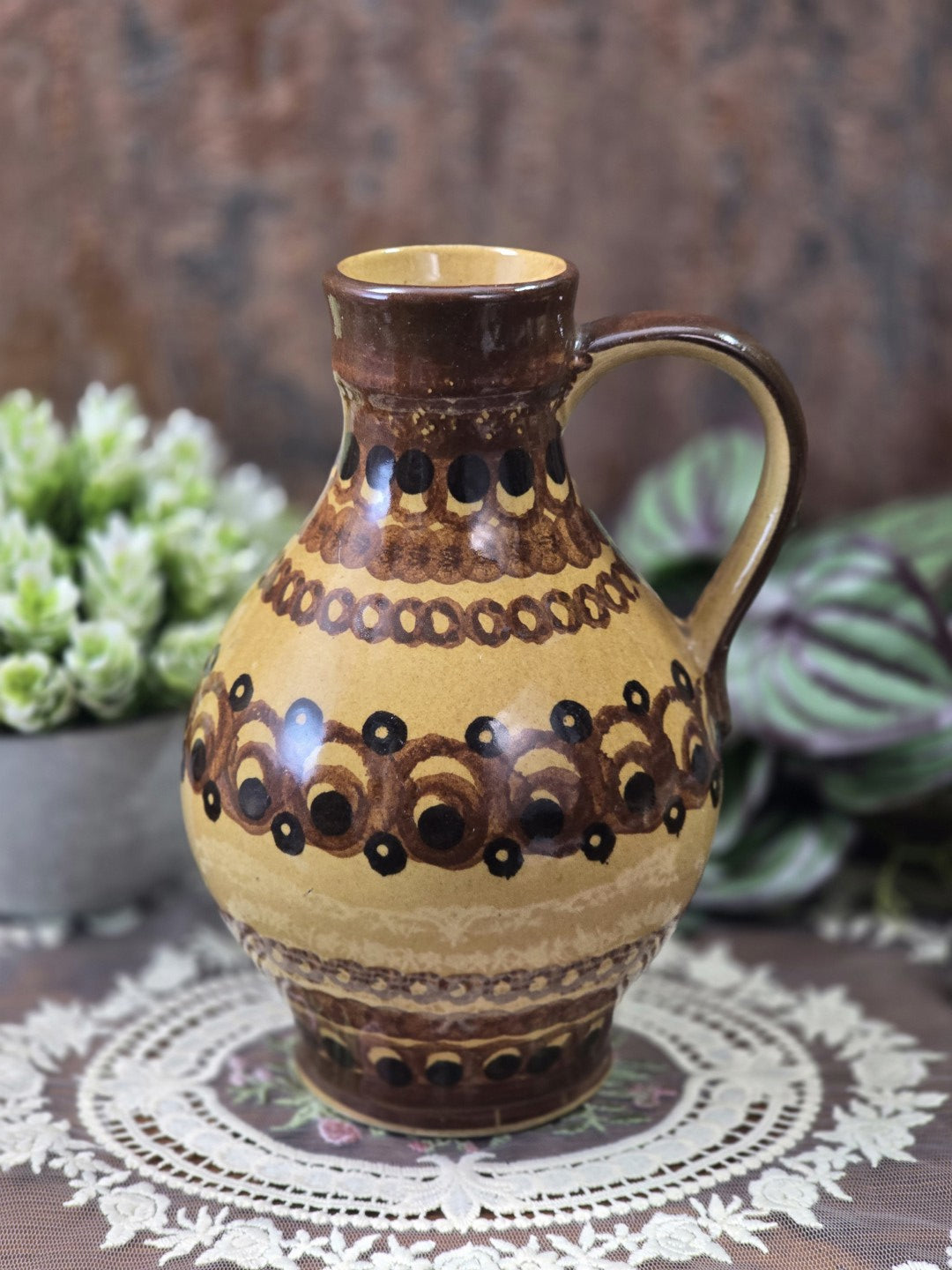 Vintage Keramik-Henkelkrug mit braunem Hals, schwarzem Punktdekor und ockergelbem Korpus vor grünen Pflanzen.