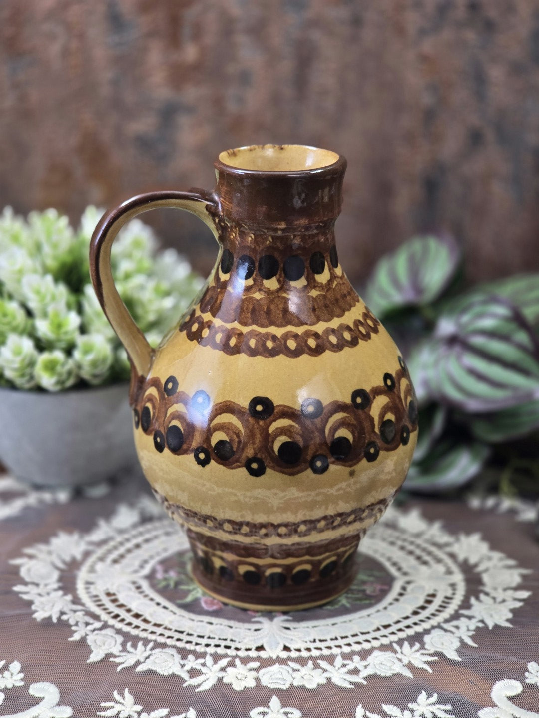 Vintage Keramikkrug mit bauchiger Form, umlaufendem Braun-Schwarz-Dekor und seitlich sichtbarem Henkel vor Pflanzenhintergrund.