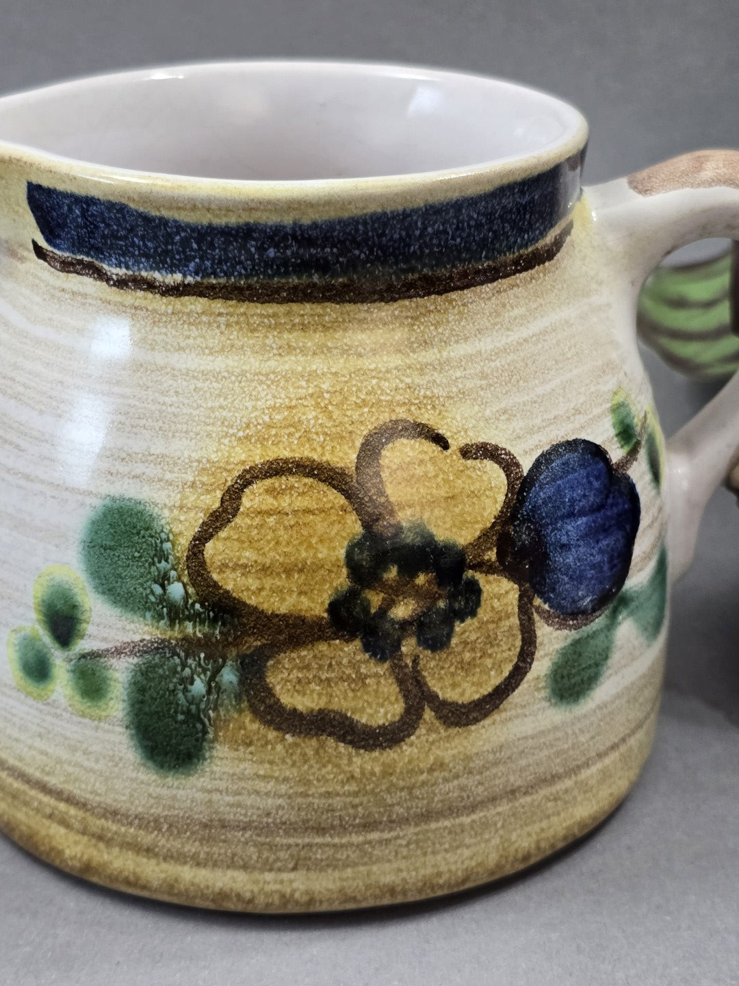 Nahaufnahme des floralen Dekors mit gelber Blume, grünen Blättern und blauem Rand auf dem Keramikkännchen