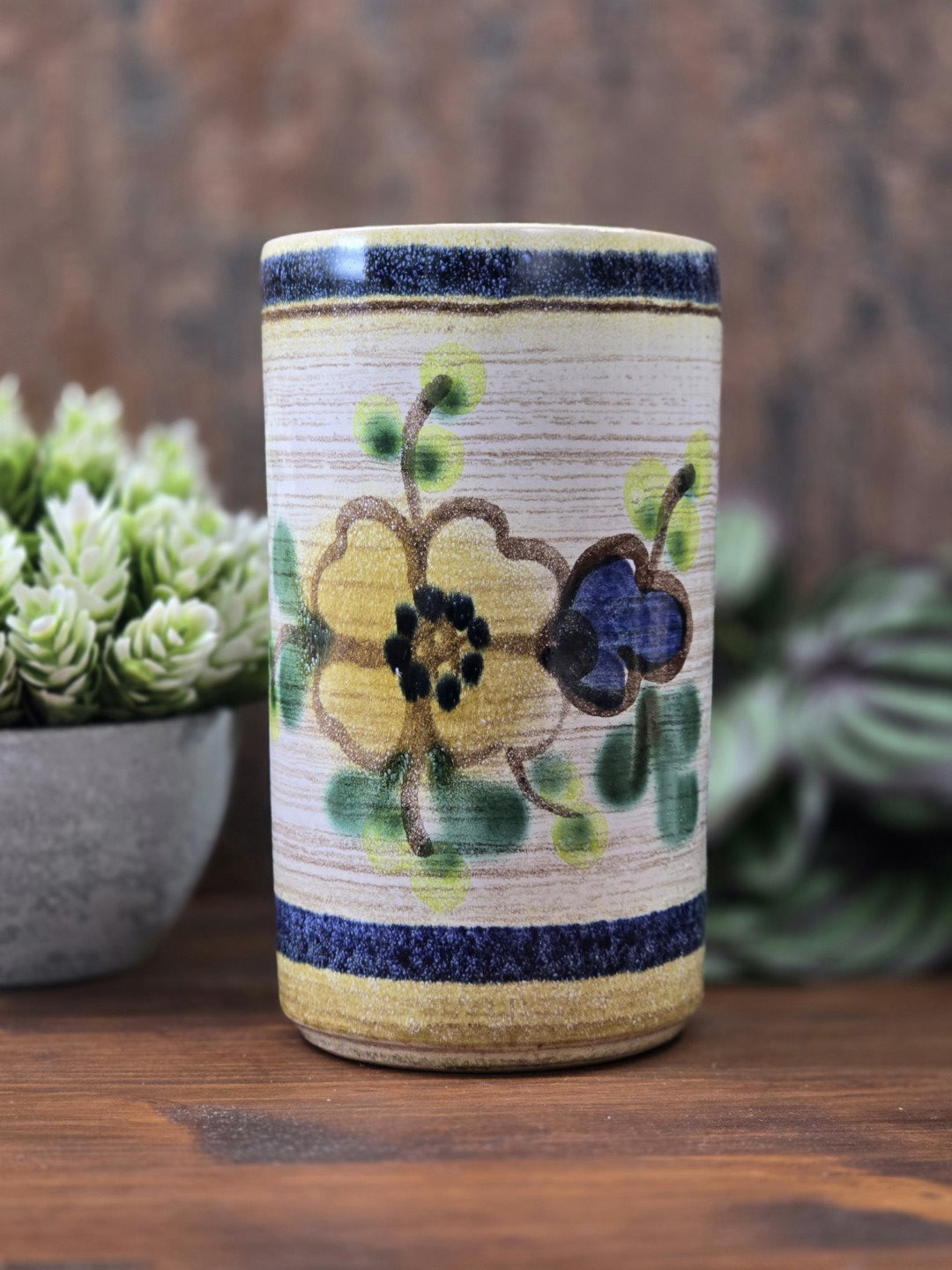 Vintage Keramikvase mit gelber und blauer Blüte, grünen Blättern, beigem Grund und dunkelblauen Randstreifen vor braunem Hintergrund.