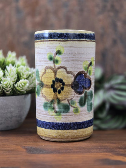 Vintage Keramikvase mit gelber und blauer Blüte, grünen Blättern, beigem Grund und dunkelblauen Randstreifen vor braunem Hintergrund.