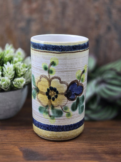 Vintage Keramikvase mit floralem Dekor in Gelb, Blau und Grün, zylindrischer Form und heller Innenwand vor Pflanzenkulisse.