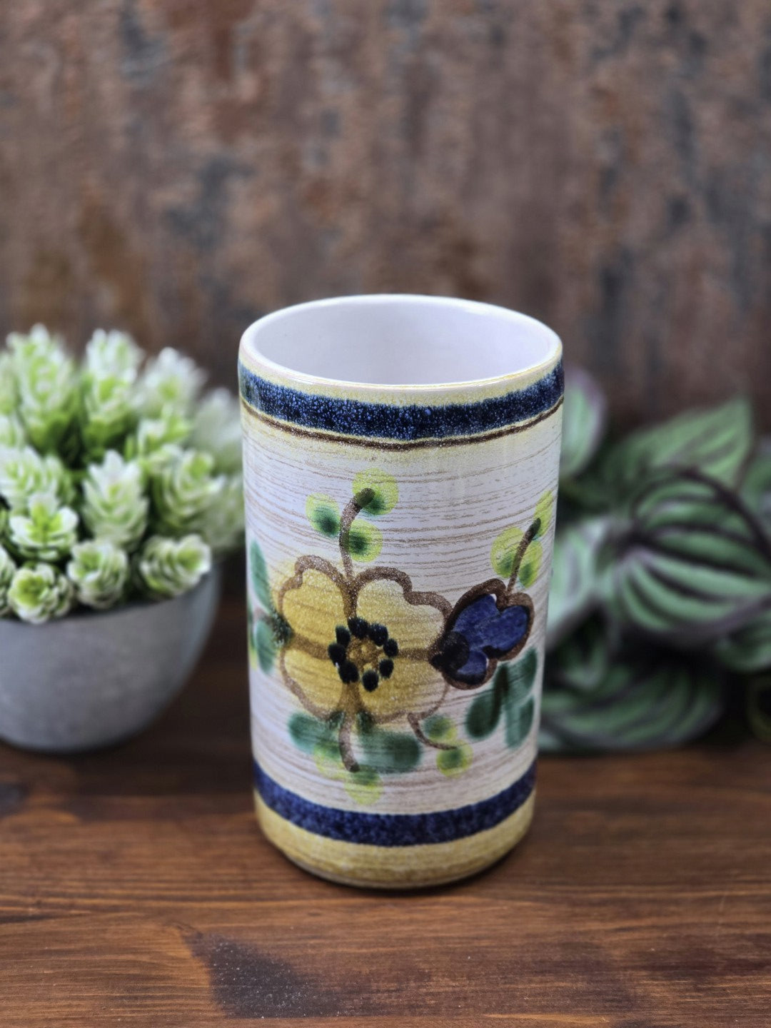 Vintage Keramikvase in zylindrischer Form mit floralem Dekor, dunkelblauen Streifen und heller Öffnung auf Holzoberfläche.