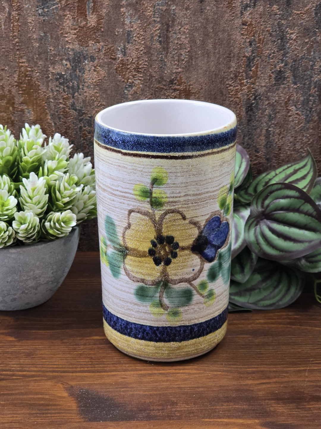Vintage Keramikvase mit gelber Blüte, blauer Blume und grünen Blättern, seitlich vor dekorativen Zimmerpflanzen fotografiert.