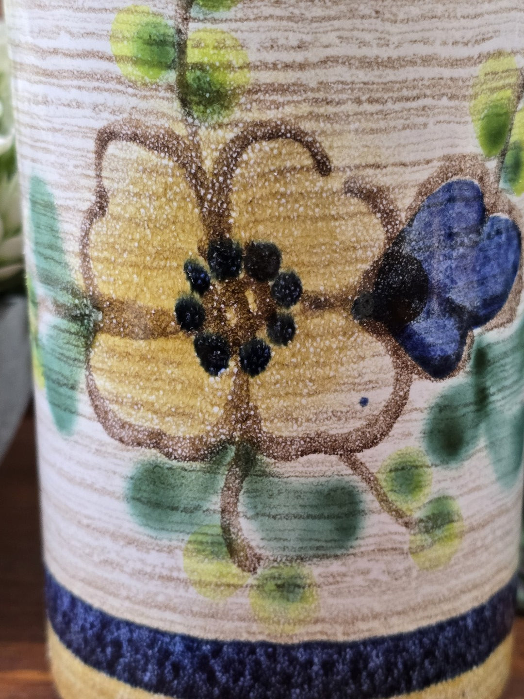 Vintage Keramikvase mit großem gelbem Blütenmotiv, blauem Blütendetail und grünen Blättern vor rustikaler Kulisse.