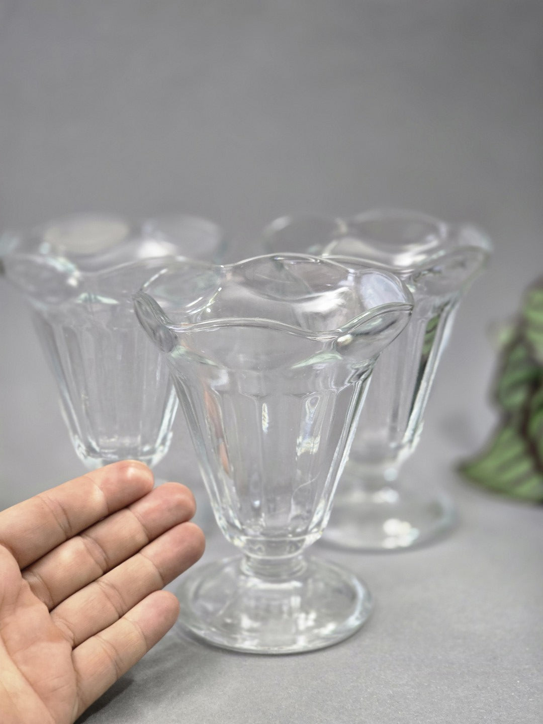 Ein transparenter Eisbecher steht im Vordergrund, dahinter sind zwei weitere Gläser unscharf zu sehen, links dient eine Hand als Größenvergleich