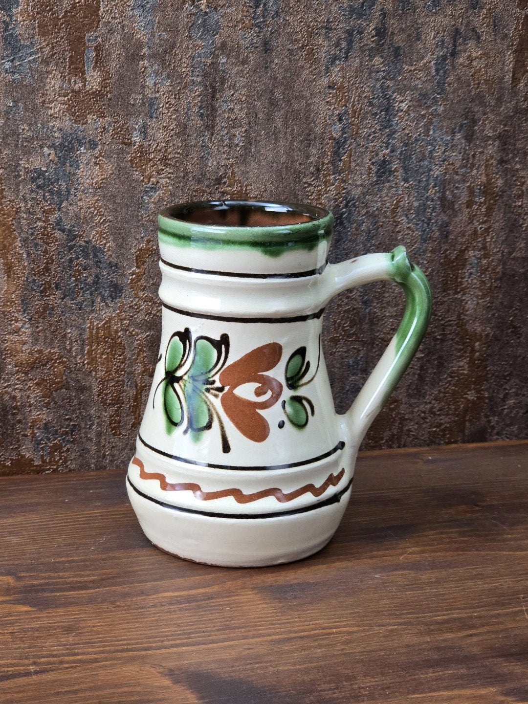 Vintage Keramikkrug mit floralem Dekor in Grün und Braun, cremefarbener Glasur, grünem Henkel und rotbrennendem Boden, dekorativ und handwerklich geprägt.