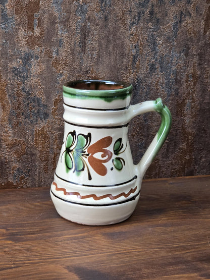 Vintage Keramikkrug mit floralem Dekor in Grün und Braun, cremefarbener Glasur, grünem Henkel und rotbrennendem Boden, dekorativ und handwerklich geprägt.