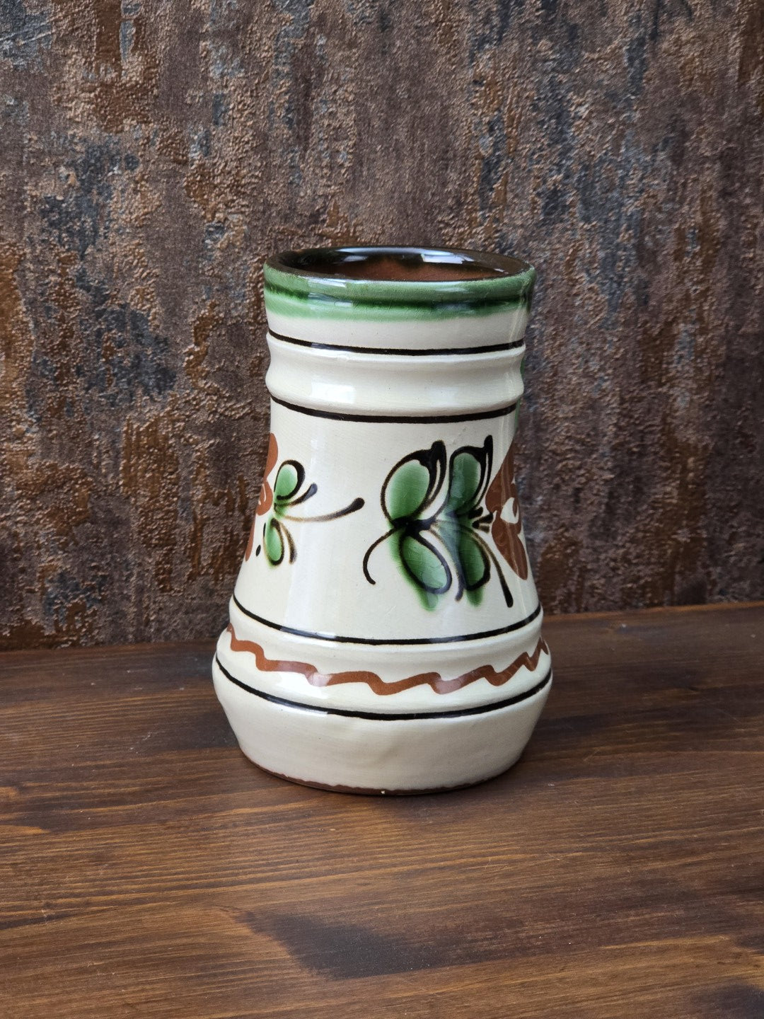 Kleiner Vintage-Krug aus glasierter Keramik mit cremefarbenem Grundton, grünem Henkel, braun-grünem Blumenmotiv und unglasiertem rötlichem Boden.