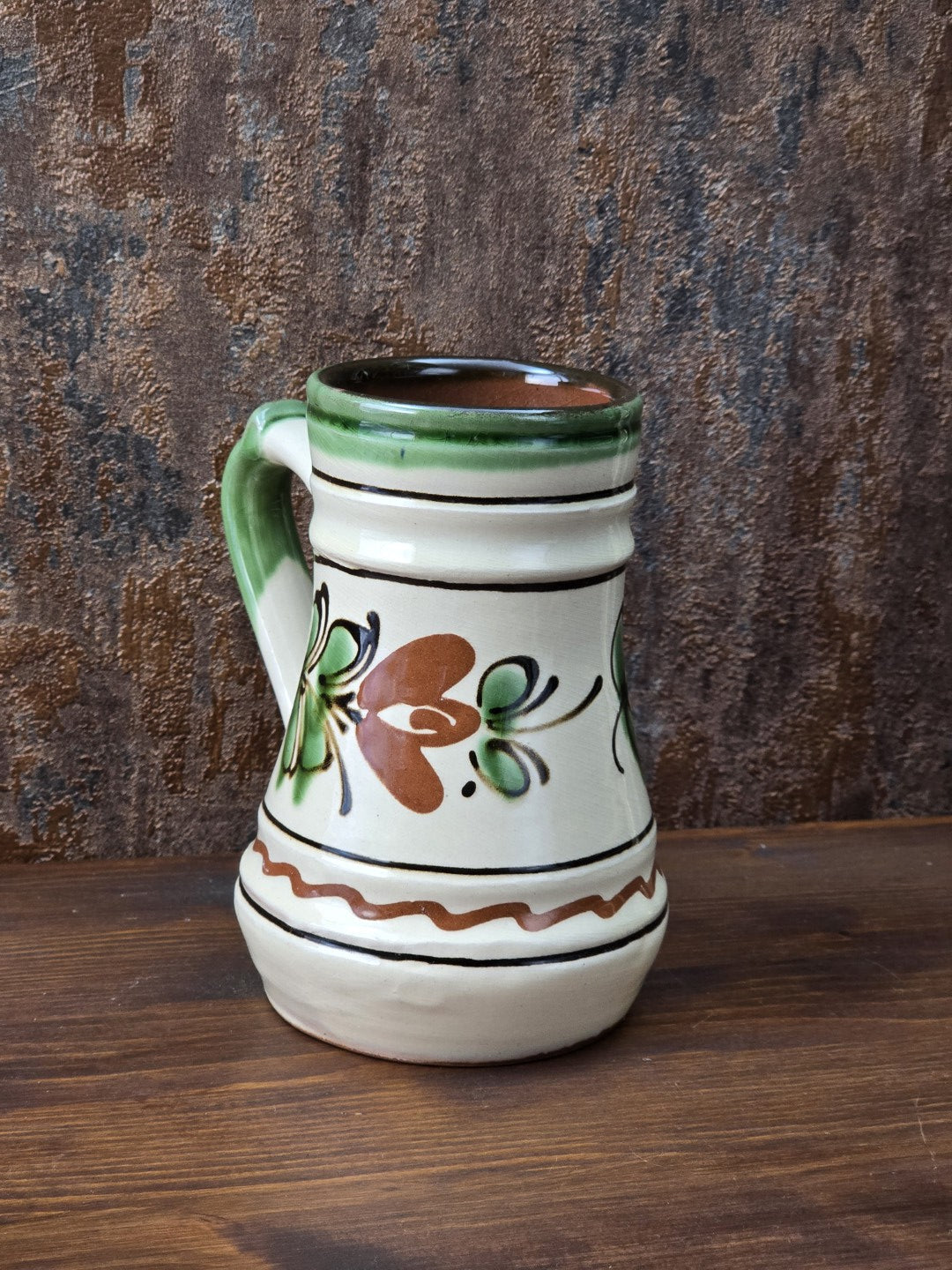 Dekorativer Keramikkrug im Vintage-Stil mit grünem Henkel, cremefarbener Glasur, rustikalem Blumendekor und rötlichem unglasiertem Standboden.