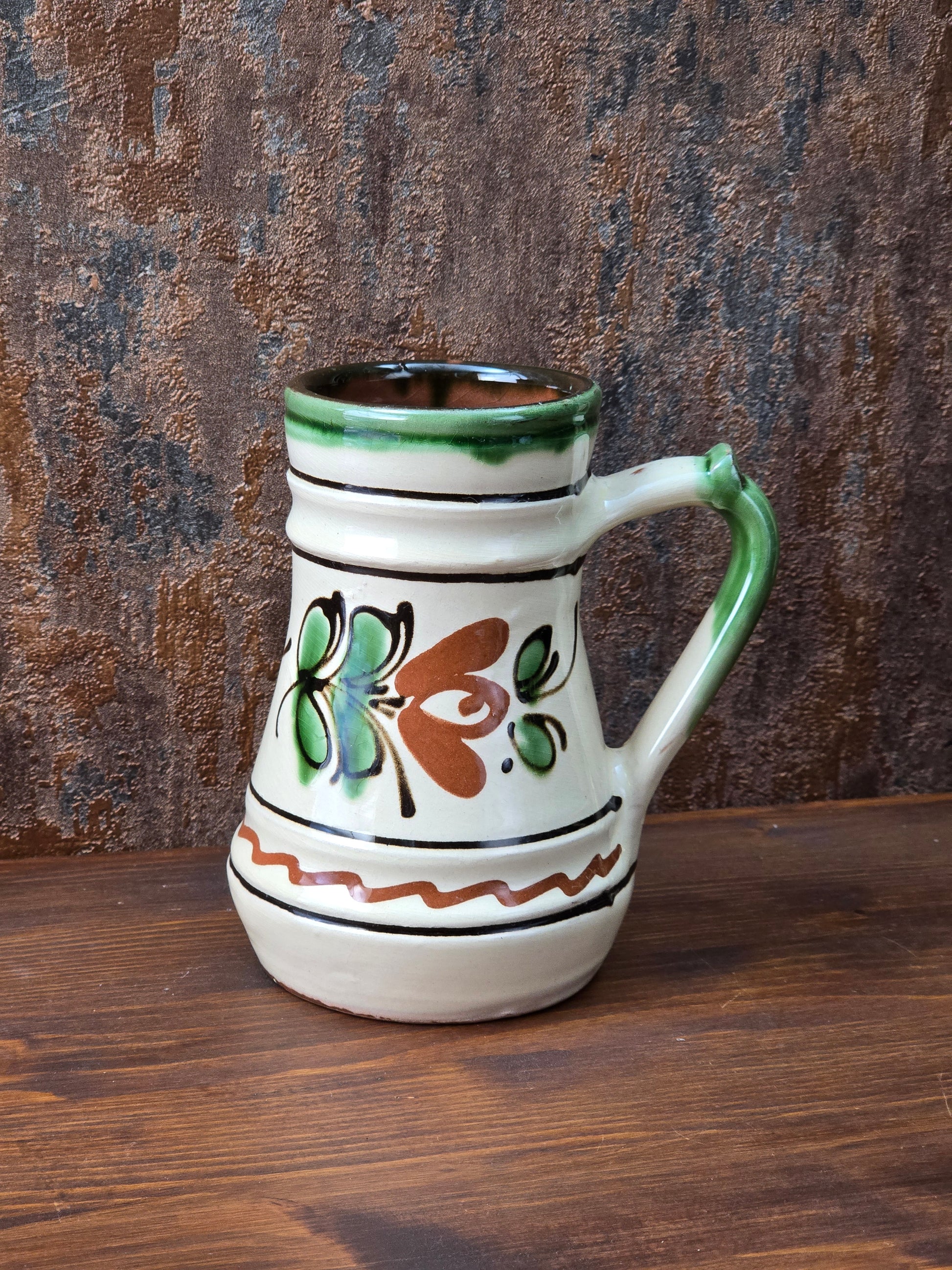 Vintage Keramikkrug mit floralem Dekor in Grün und Braun, cremefarbener Glasur, grünem Henkel und sichtbarem rotbrennendem Keramikscherben am Boden.