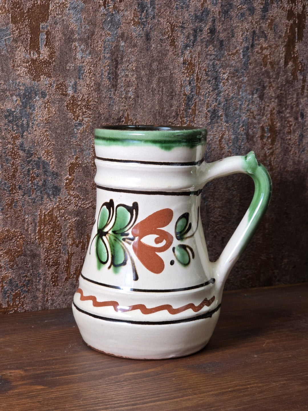 Vintage Zierkrug aus Keramik mit grün-braunem Blumendekor, heller Glasur, grünem Henkel und handwerklich wirkendem rotbrennendem Boden.
