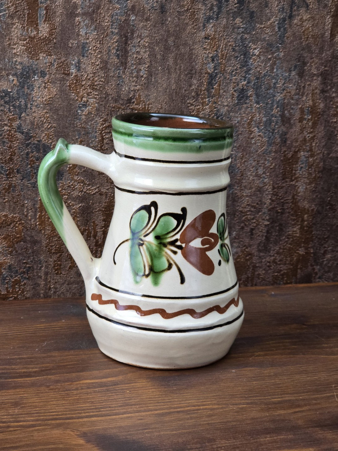 Rustikaler Vintage Keramikkrug mit cremefarbener Glasur, grünem Henkel, floraler Bemalung in Braun und Grün und sichtbarem rötlichem Boden.