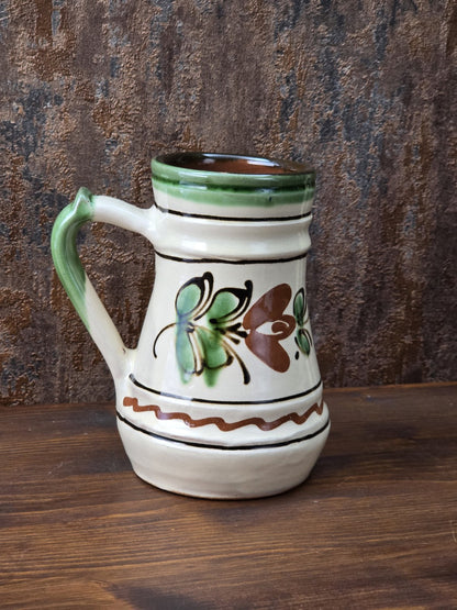 Rustikaler Vintage Keramikkrug mit cremefarbener Glasur, grünem Henkel, floraler Bemalung in Braun und Grün und sichtbarem rötlichem Boden.