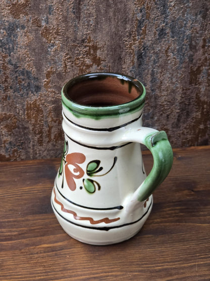 Vintage Keramik Krug mit handwerklichem Charakter, grünem Henkel, hellem cremefarbenem Glasurton und floralem Dekor in Braun und Grün.