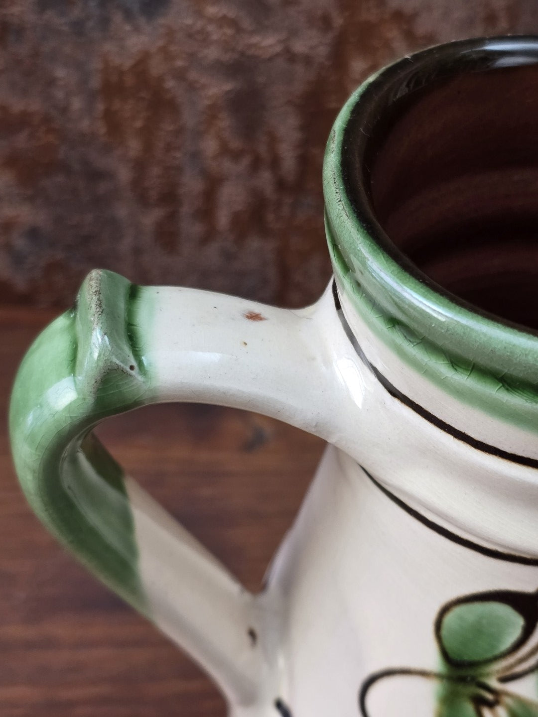 Handwerklich geprägter Vintage Keramikkrug mit grünem Henkel, cremefarben glasierter Oberfläche, Blumenmotiv in Grün und Braun und rotbrennendem Boden.