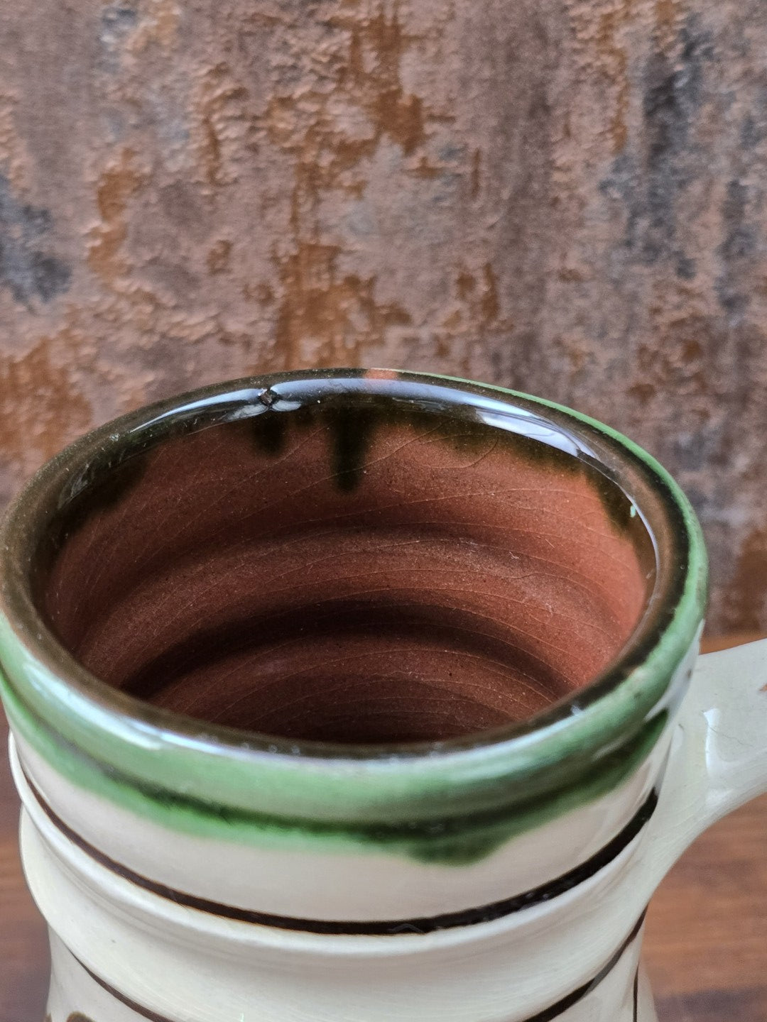Vintage Henkelkrug mit floraler Bemalung in Grün und Braun, cremefarbener Glasur, grünem Akzenthenkel und rotbrennendem Keramikscherben.