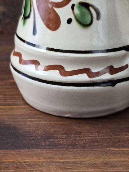Kleiner Keramik-Zierkrug mit cremefarbener Glasur, grünem Henkel, handgemalt wirkendem Blumendekor und rötlich gebranntem Boden.