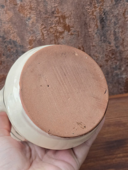 Rustikaler Keramik Krug im Vintage-Stil mit grünem Henkel, floralem Dekor in Braun und Grün, cremefarbener Glasur und sichtbarem Terrakotta-Boden.