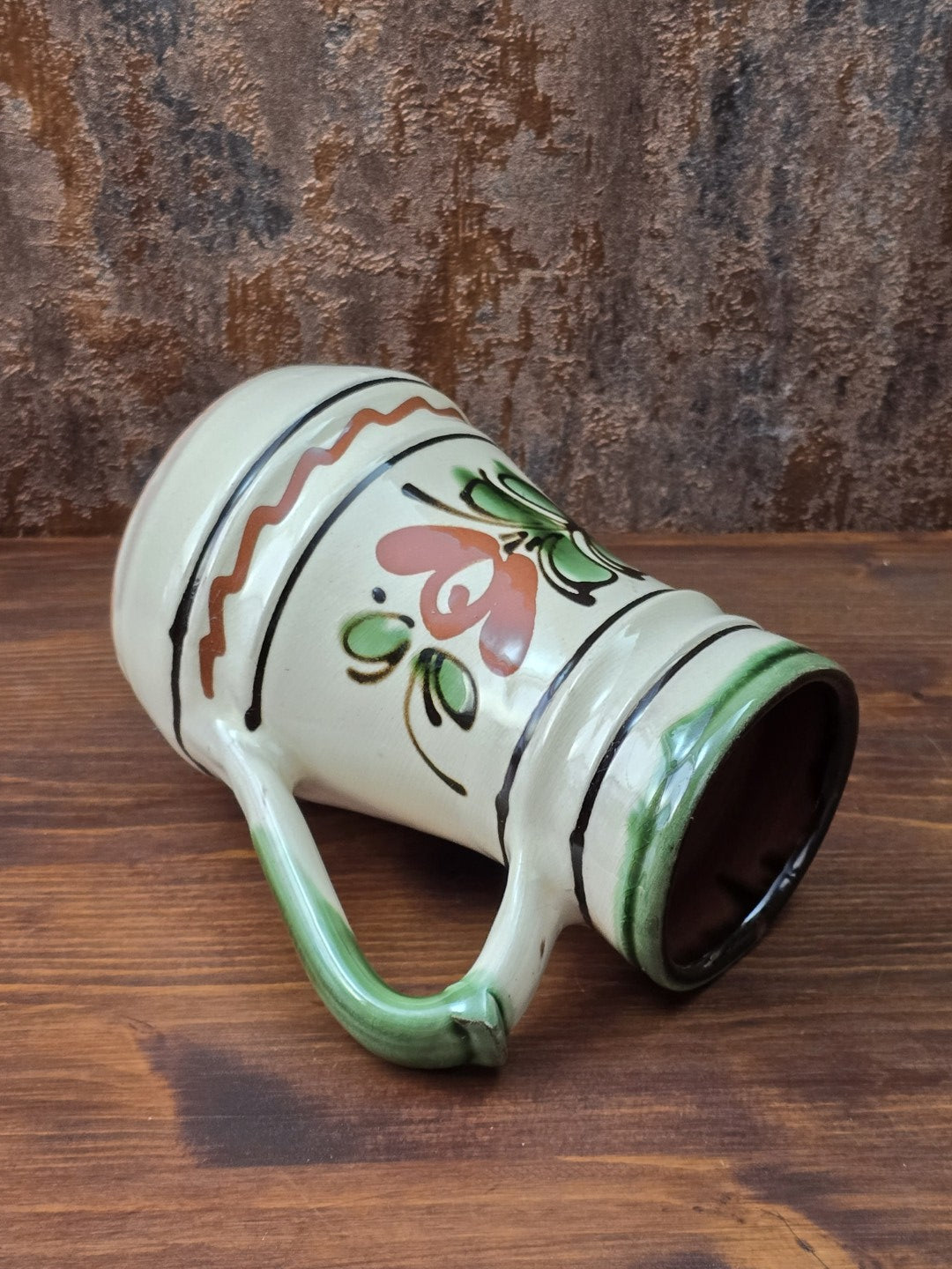 Vintage Keramikkrug mit hellem cremefarbenem Glasurgrund, grünem Henkel, braun-grünem Blumenmuster und handwerklich geprägter Oberfläche.