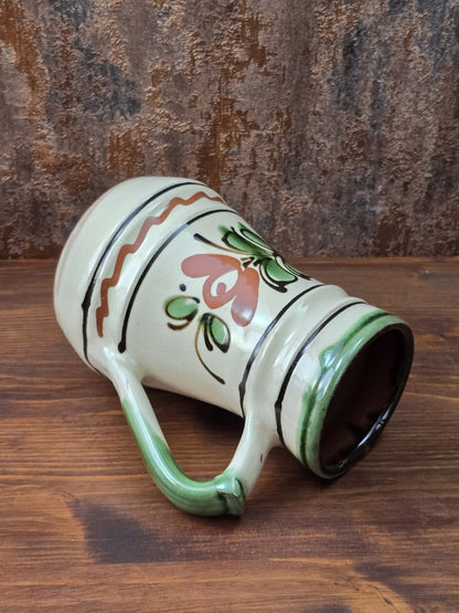 Vintage Keramikkrug mit hellem cremefarbenem Glasurgrund, grünem Henkel, braun-grünem Blumenmuster und handwerklich geprägter Oberfläche.