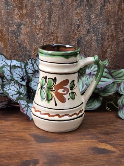 Dekorativer kleiner Vintage-Krug mit grünem Henkel, cremefarbener Keramikglasur, floralem Grün-Braun-Dekor und rotbrennendem unglasiertem Boden.