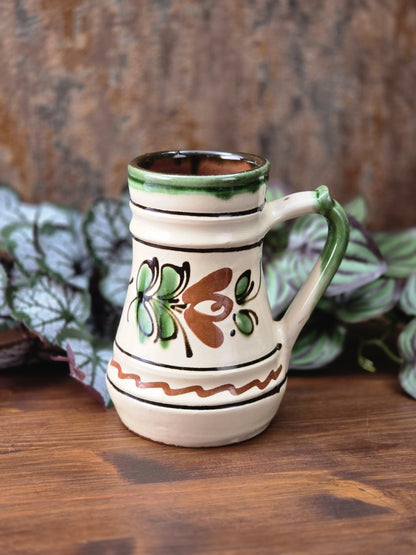 Vintage Zierkrug aus glasierter Keramik mit cremefarbenem Grundton, grünem Henkel, rustikalem Floraldekor und sichtbarem rotbraunem Boden.