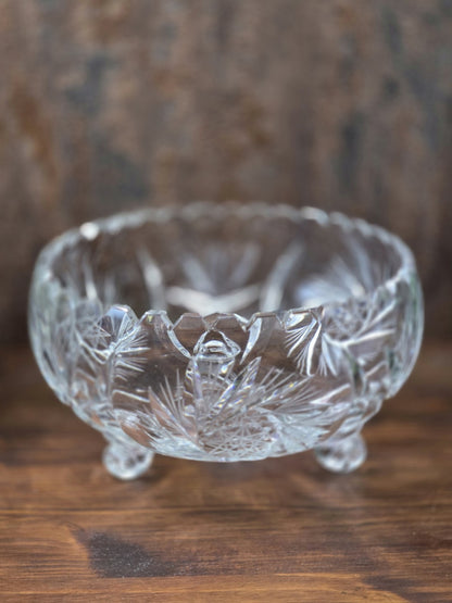 Vintage Kristallglasschale auf drei Füßen mit facettiertem Rand und floralem Sternschliff, frontal auf dunkler Holzfläche fotografiert.
