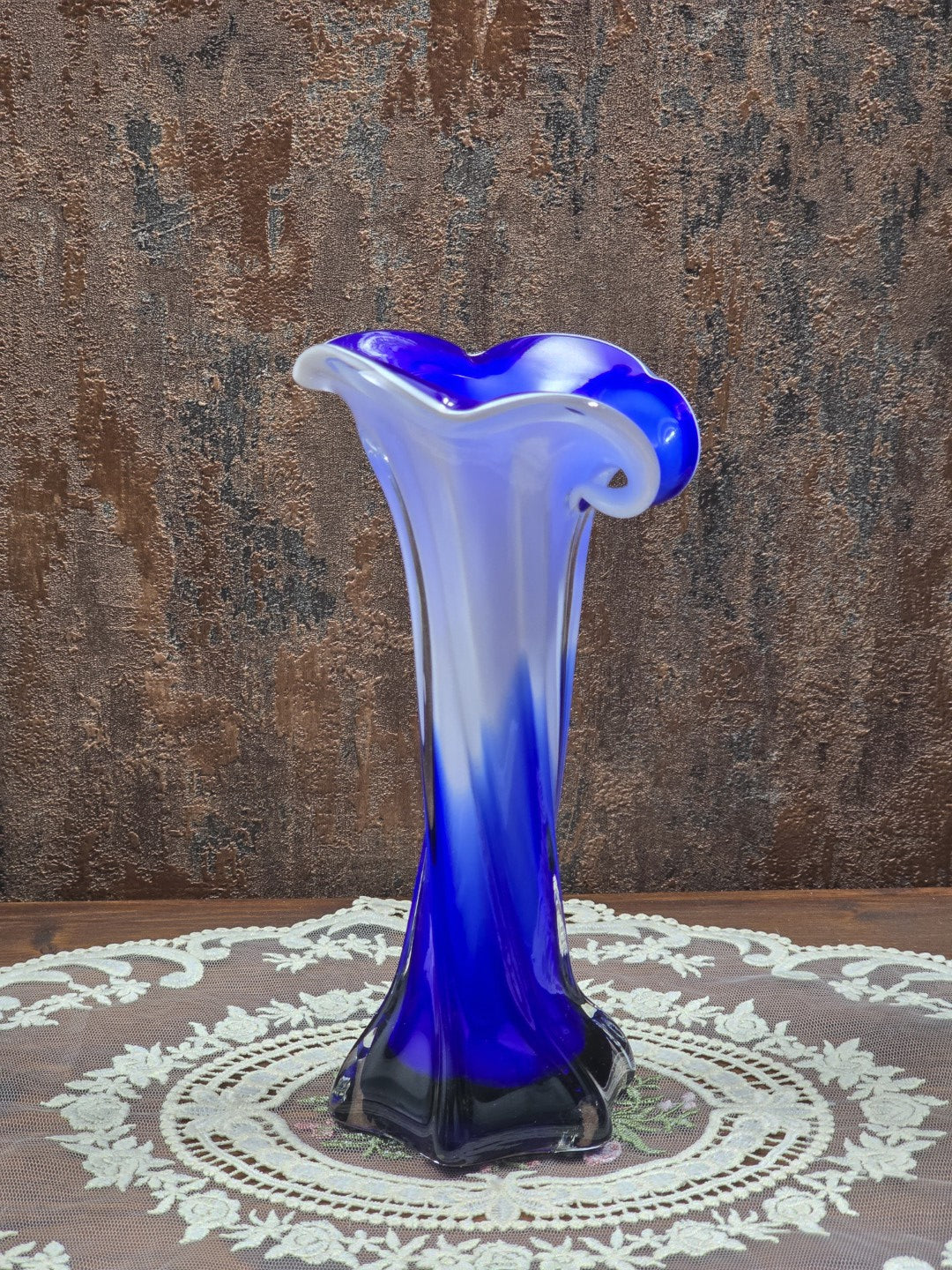 Glasvase mit blau-weißem Verlauf, transparentem Sternfuß und geschwungener Mündung zwischen grünen Blattpflanzen