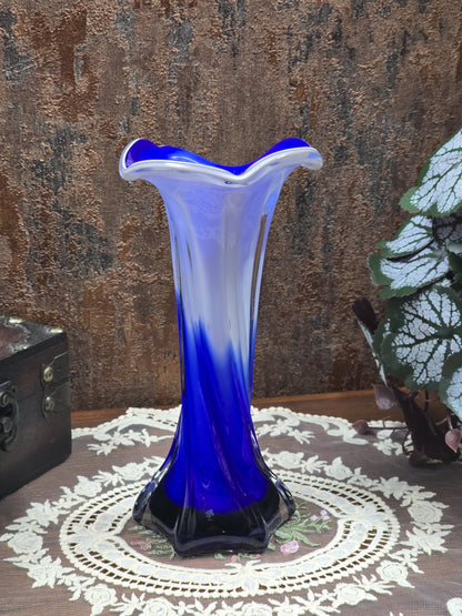 Glasvase mit blauem Verlauf, weißem Glas und seitlich geschwungener Mündung neben Blattpflanze und kleiner Truhe