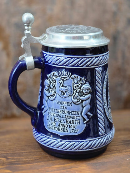 Vintage Marzi & Remy Bierkrug mit Zinndeckel, blauem Henkel und Relief des Eisenbarth-Wappens samt Inschrift in Seitenansicht.