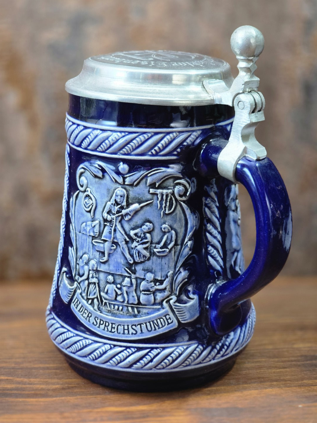 Vintage Steinzeug-Bierkrug mit kobaltblauer Glasur, Zinndeckel und plastischem Relief „In der Sprechstunde“ in schräger Seitenansicht.