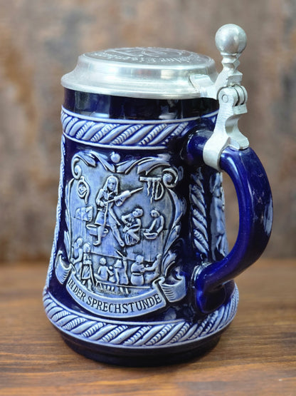 Vintage Steinzeug-Bierkrug mit kobaltblauer Glasur, Zinndeckel und plastischem Relief „In der Sprechstunde“ in schräger Seitenansicht.