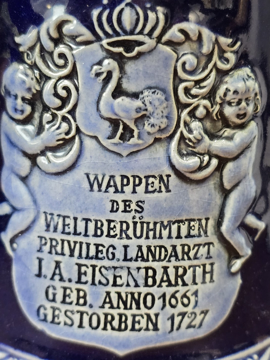 Vintage Bierkrug mit Eisenbarth-Wappen, zwei plastischen Figuren und beschriftetem Relief zu Johann Andreas Eisenbarth in Nahaufnahme.