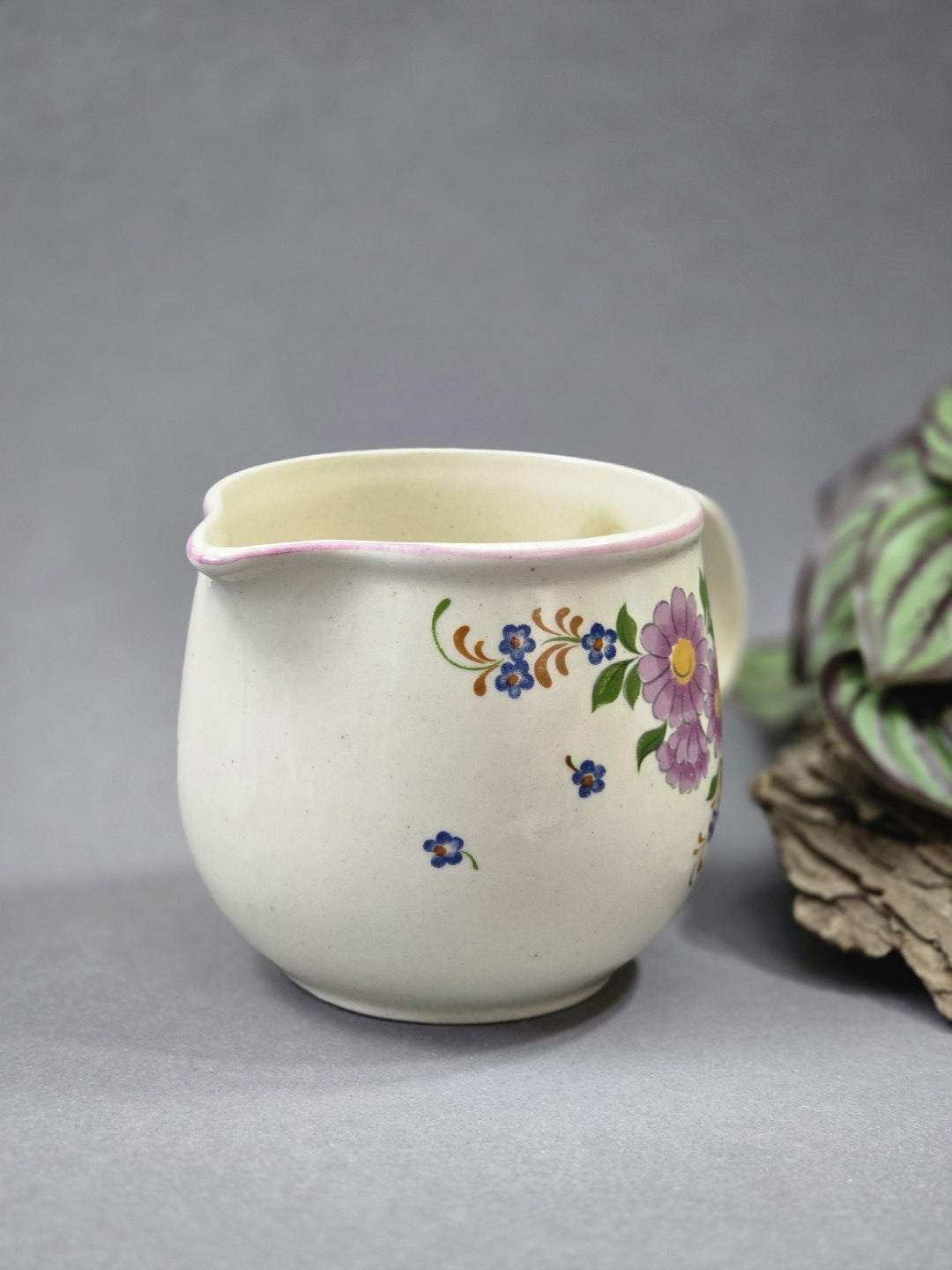 Cremefarbenes Keramik Milchkännchen von der Seite, mit zartem rosa Rand, kleinen blauen Blüten und angeschnittenem lila Blumenmotiv.