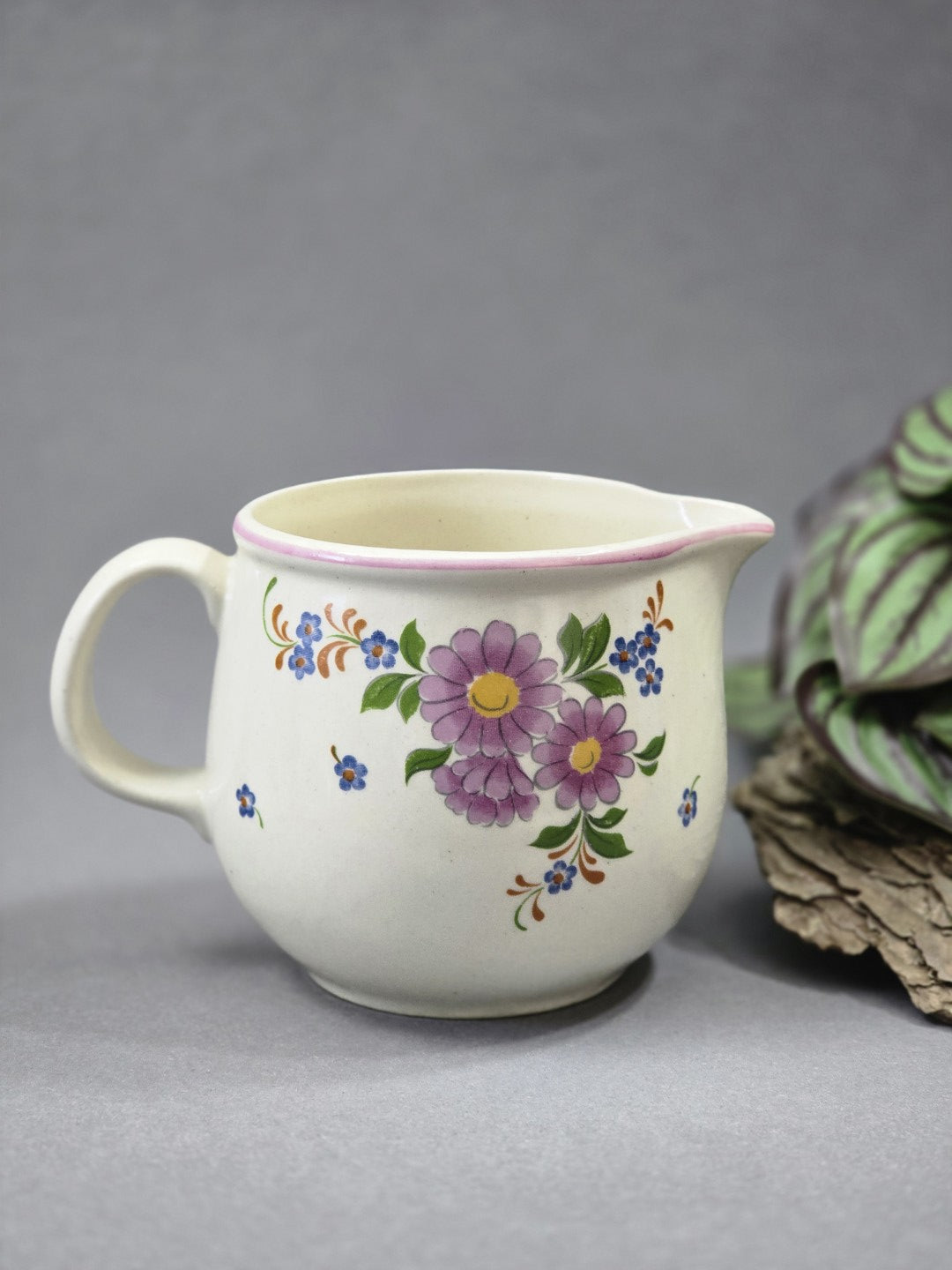 Frontansicht eines hellen Milchkännchens mit floraler Verzierung, rundem Henkel links, Ausguss rechts und weicher bauchiger Form.