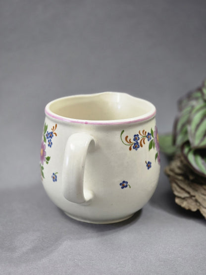 Rückseite des cremefarbenen Milchkännchens mit Henkel, kleinen blauen Blüten, zartem rosa Rand und glatter heller Keramikglasur.
