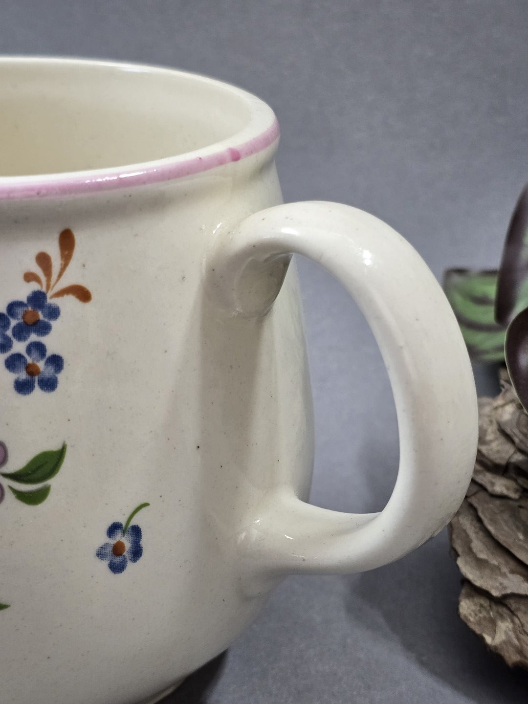 Nahaufnahme des runden Henkels mit heller Glasur, daneben kleine blaue Blüten und der rosa Rand des Vintage Milchkännchens.