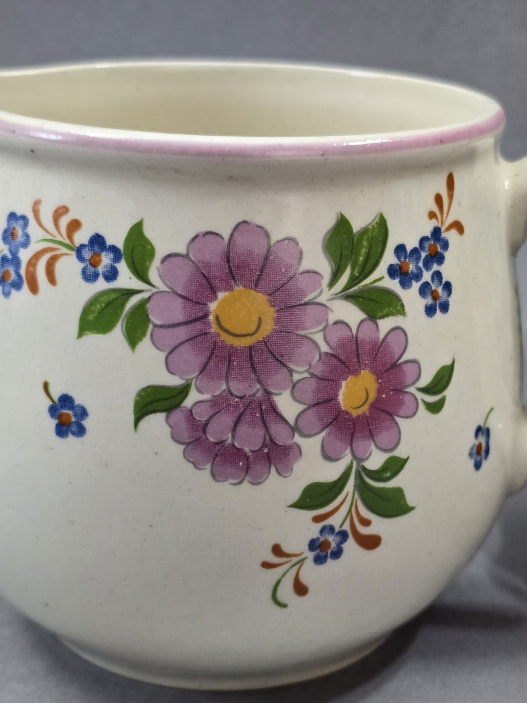 Florales Detail auf hellem Keramikkorpus mit lila Blüten, blauen Blumen, grünen Blättern und gelben Blütenmitten.